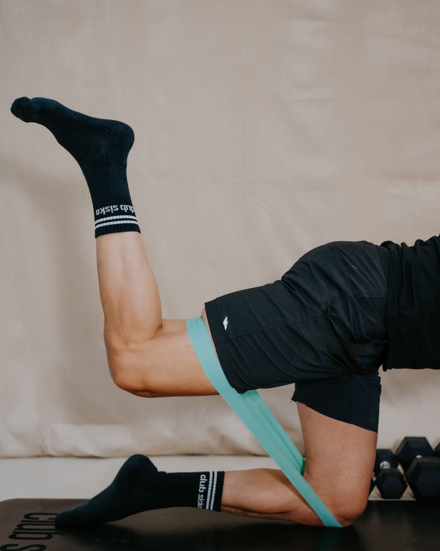 Extension de jambe à quatre pattes avec élastique, sur un tapis Club Sisko, haltères en arrière-plan